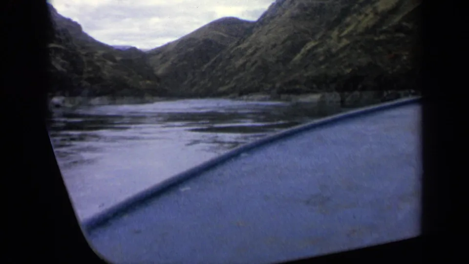 Boat-side perspective on an Idaho river in 1963, with muted hillsides and calm water under overcast light.