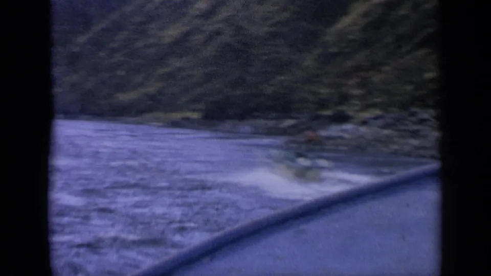 A distant paddler appears on the Idaho river near small waves, seen from a moving boat in 1963 home-movie footage.