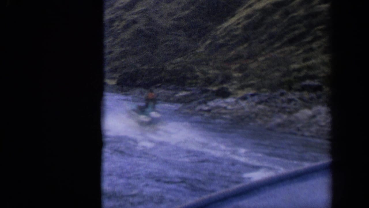 Small motorized craft speeds across water near a rocky shore and steep hillside.
