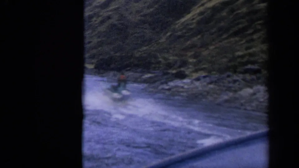 1963 Idaho 8mm frame of a distant rider on a small motorized craft crossing choppy water near a rugged shoreline.
