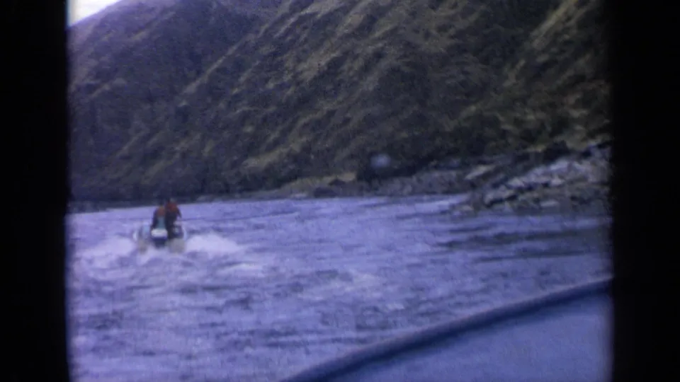 The small watercraft rider remains distant in a 1963 Idaho home-movie view of choppy water, wake, rocks, and steep hillsides.