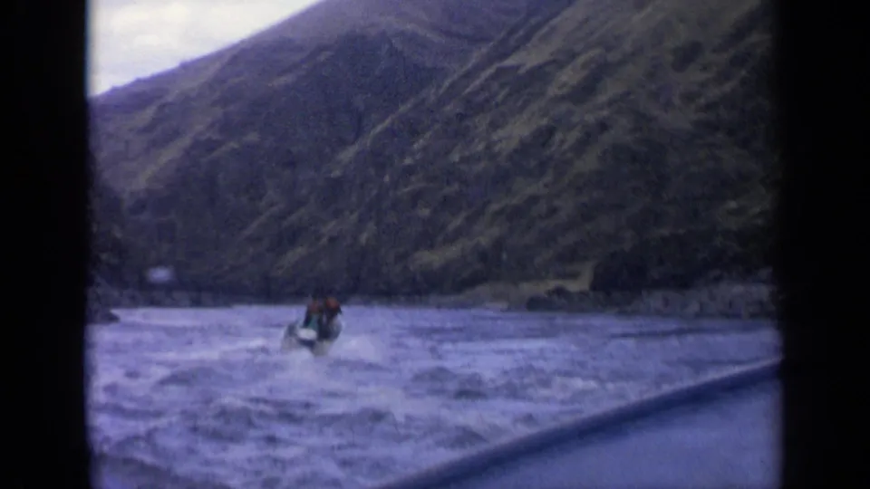 Home-movie frame of a small motorboat on a turbulent river beneath steep Idaho mountain terrain.