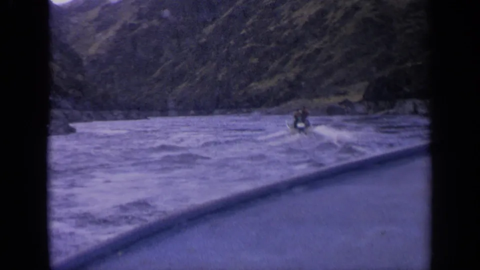 A small motorboat travels away across rough Idaho lake water, with a rocky shoreline and steep hillside in the background.