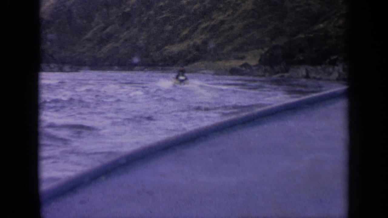 Small boat on a rushing river below steep mountain slopes, seen through dark 8mm frame borders.