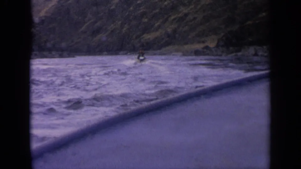 1963 Idaho home-movie frame with a small boat on choppy river water near steep mountain terrain.