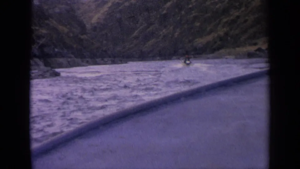 1963 Idaho 8mm frame showing a distant boat moving through fast water in a mountain river canyon.