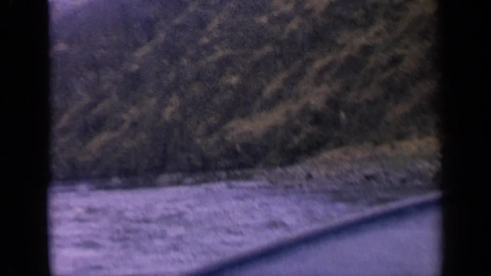 1963 Idaho 8mm view of a river beside a rocky bank and forested mountain slope, framed by dark film gate edges.