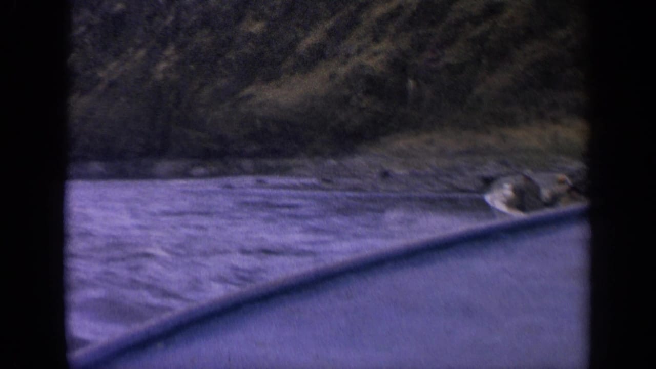 1963 Idaho 8mm frame with a small passenger boat at the right edge of a river below wooded mountain slopes.