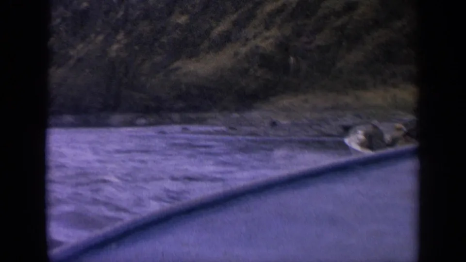 1963 Idaho 8mm frame with a small passenger boat at the right edge of a river below wooded mountain slopes.
