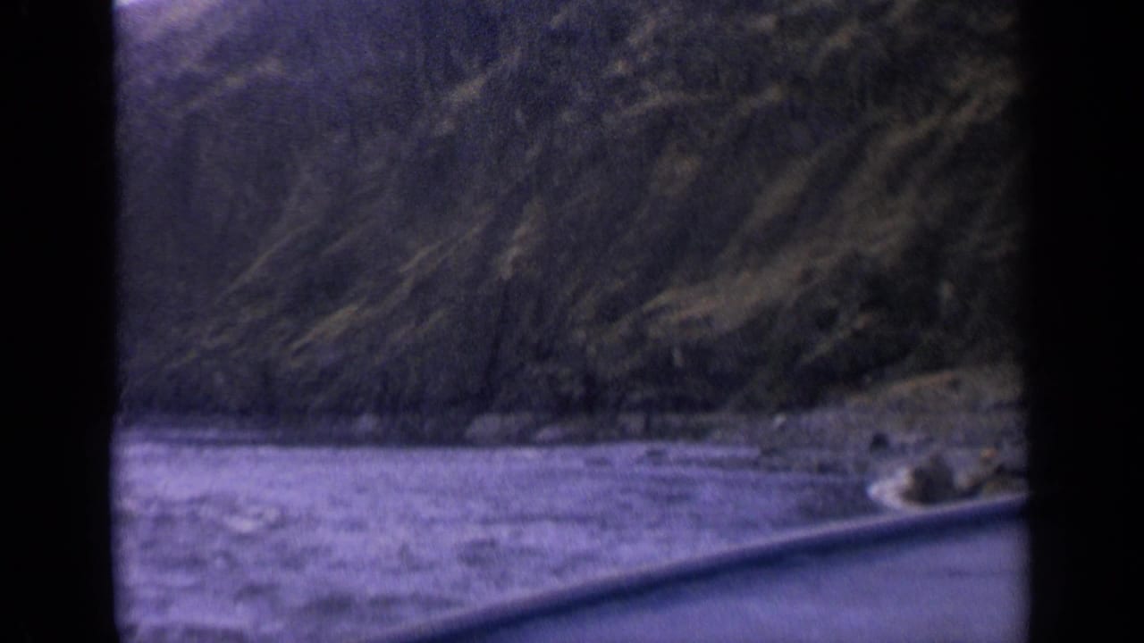 Choppy water and a distant small boat near a steep wooded shoreline, partly framed by dark film edges.