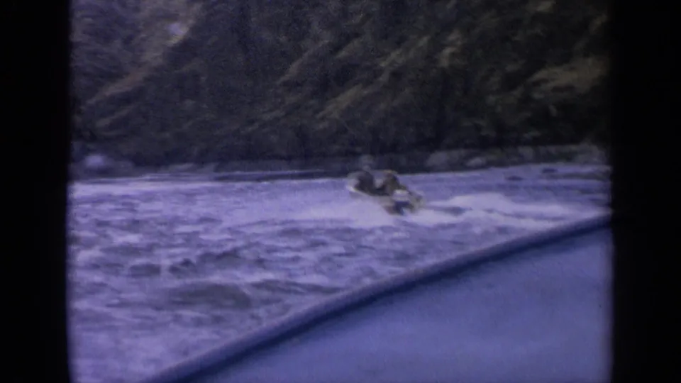 1963 Idaho 8mm frame showing a small speedboat and faint occupants speeding over rough water, filmed from behind a dark foreground edge.
