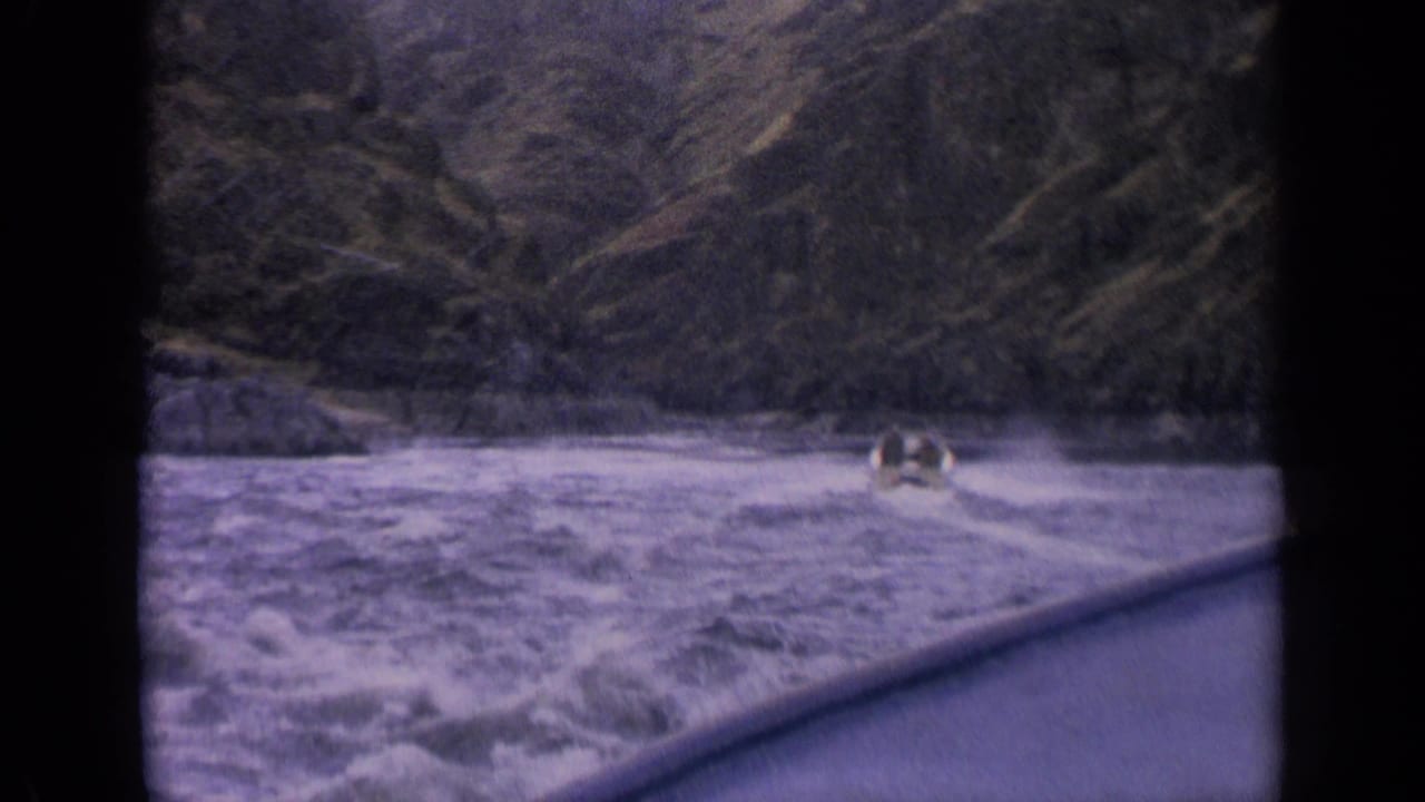 1963 home-movie frame from Idaho showing a small boat surrounded by frothy rapids and a dark wooded mountain backdrop.
