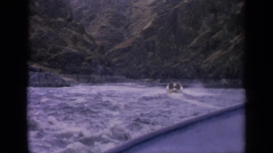 1963 home-movie frame from Idaho showing a small boat surrounded by frothy rapids and a dark wooded mountain backdrop.