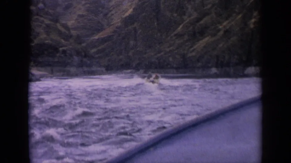 Small occupied boat continues through rough Idaho mountain river water, framed by dark 8mm borders and purple color shift.