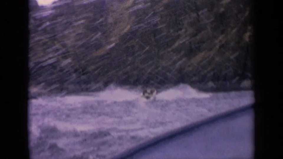 1963 Idaho 8mm frame of a small speedboat crossing a lake, with white spray against a steep wooded shoreline.