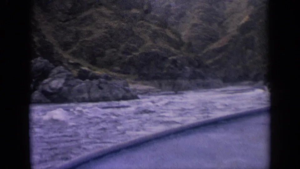 1963 Idaho lake view from a moving boat, showing water, rocky shore, and steep wooded hills.