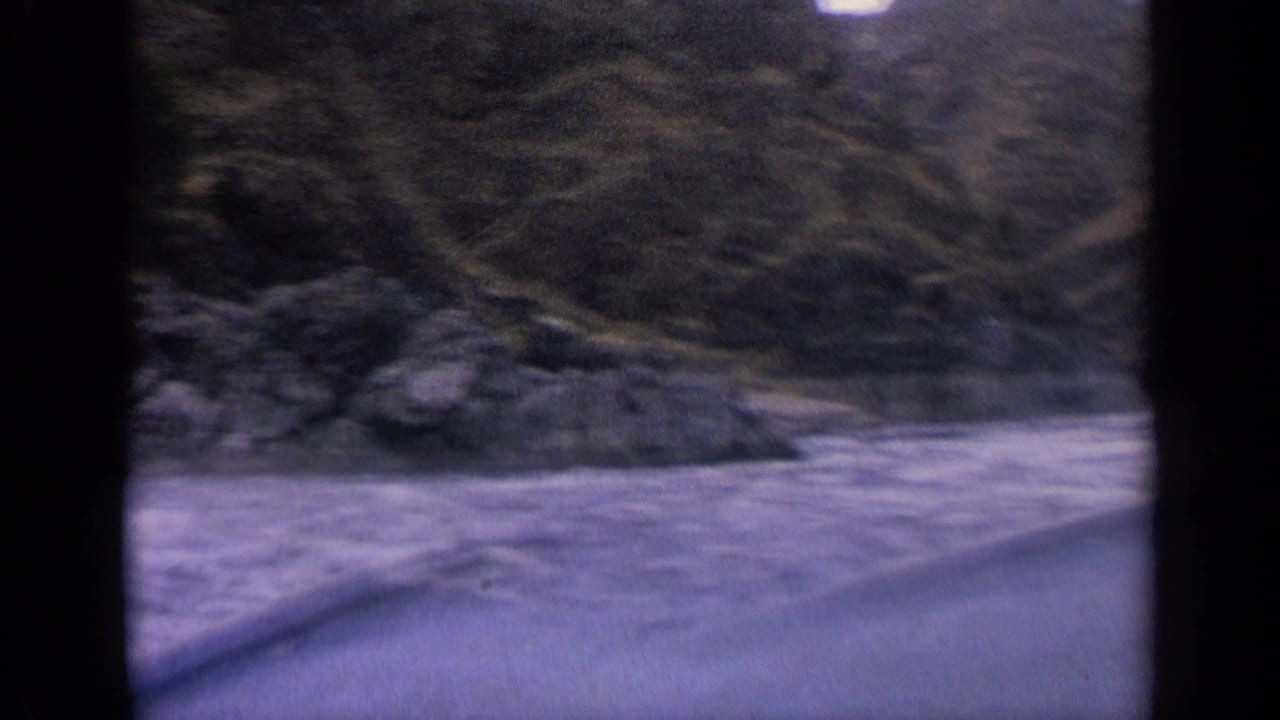 1963 Idaho 8mm frame showing blue river water from a moving boat, with dark trees and a sloped bank beyond.