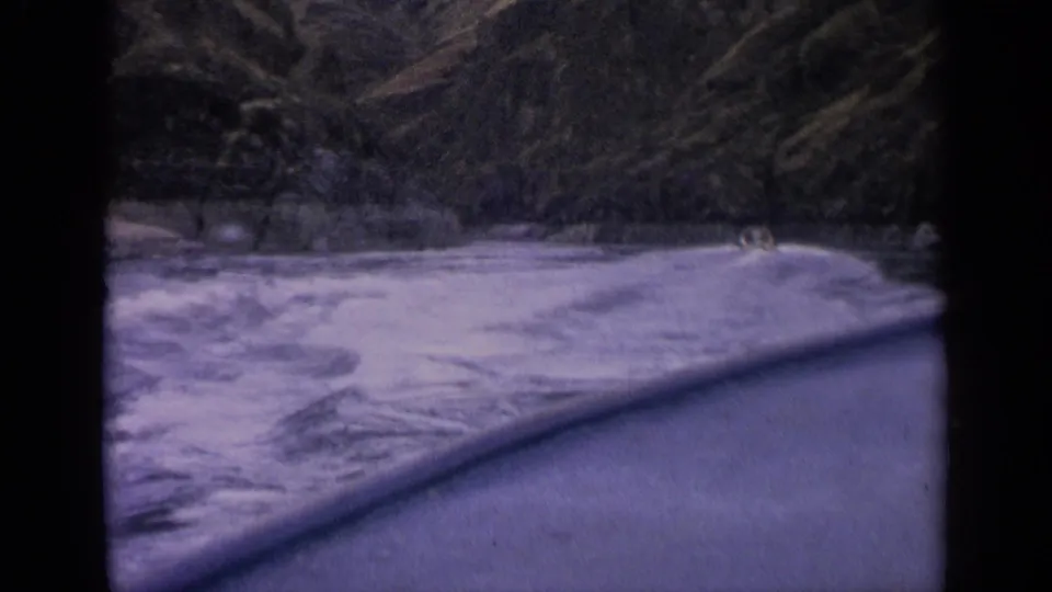 1963 Idaho river view from a boat, with foamy blue water, a curved rail or edge, and dense forest on the bank.
