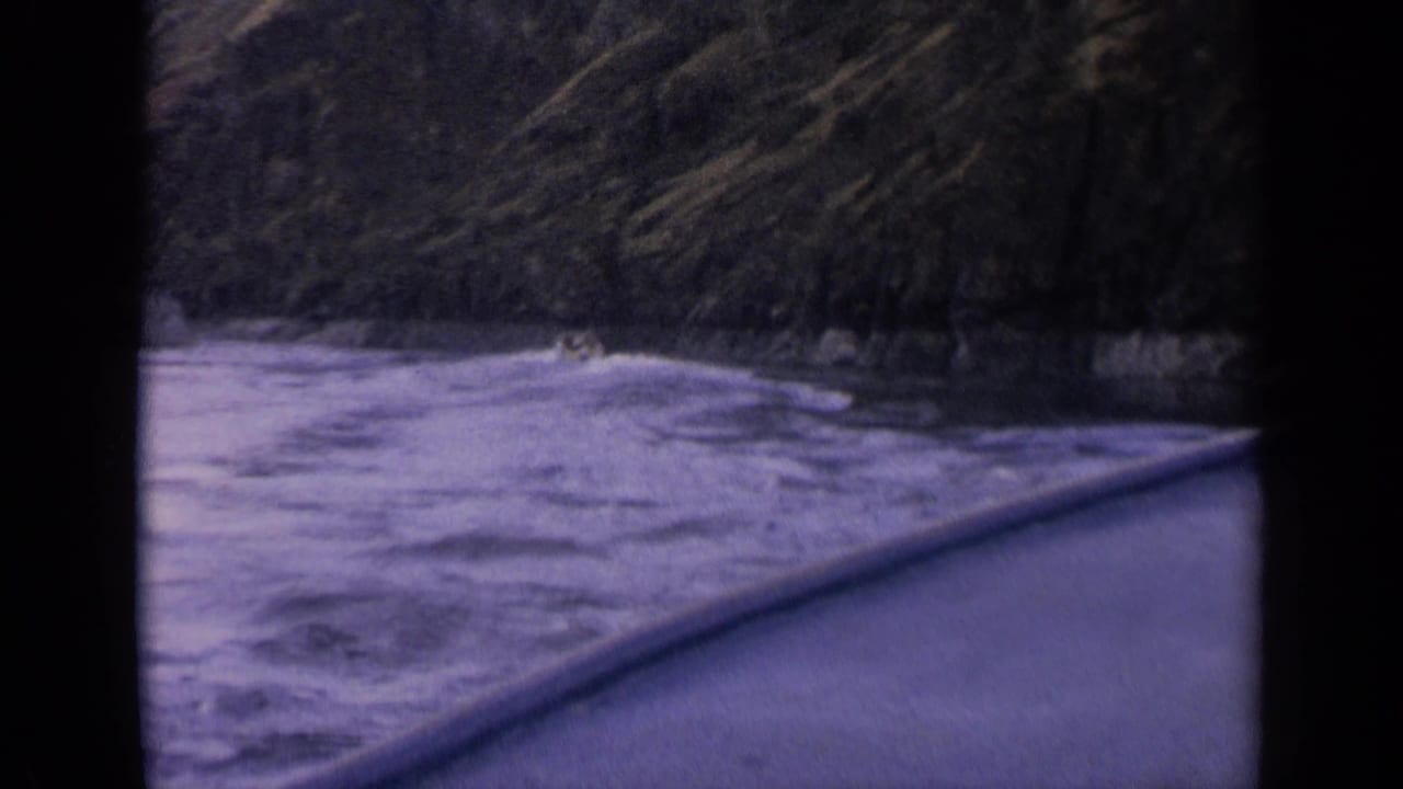 1963 Idaho 8mm still with a low passenger view over river wake, boat edge, and a dark tree-lined shore.
