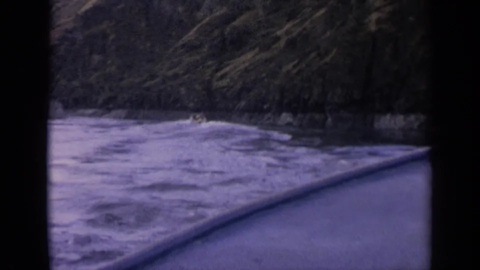 1963 Idaho 8mm still with a low passenger view over river wake, boat edge, and a dark tree-lined shore.