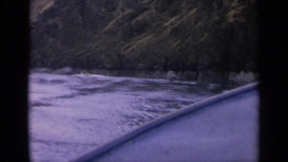 First-person 8mm view from a canoe passing through choppy Idaho river water near a wooded rock bank.