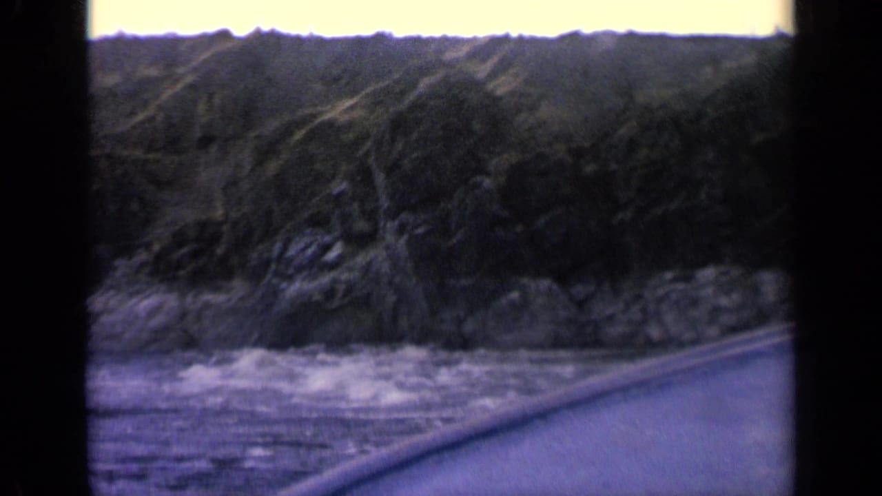Home-movie frame showing the canoe bow beside rough water under a dark wooded bank in Idaho river terrain.