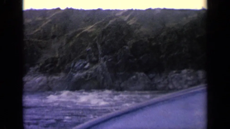 Home-movie frame showing the canoe bow beside rough water under a dark wooded bank in Idaho river terrain.