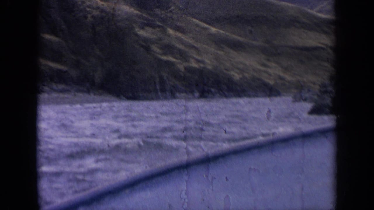 1963 Idaho 8mm frame showing a river below steep rocky hills from a moving roadside perspective.