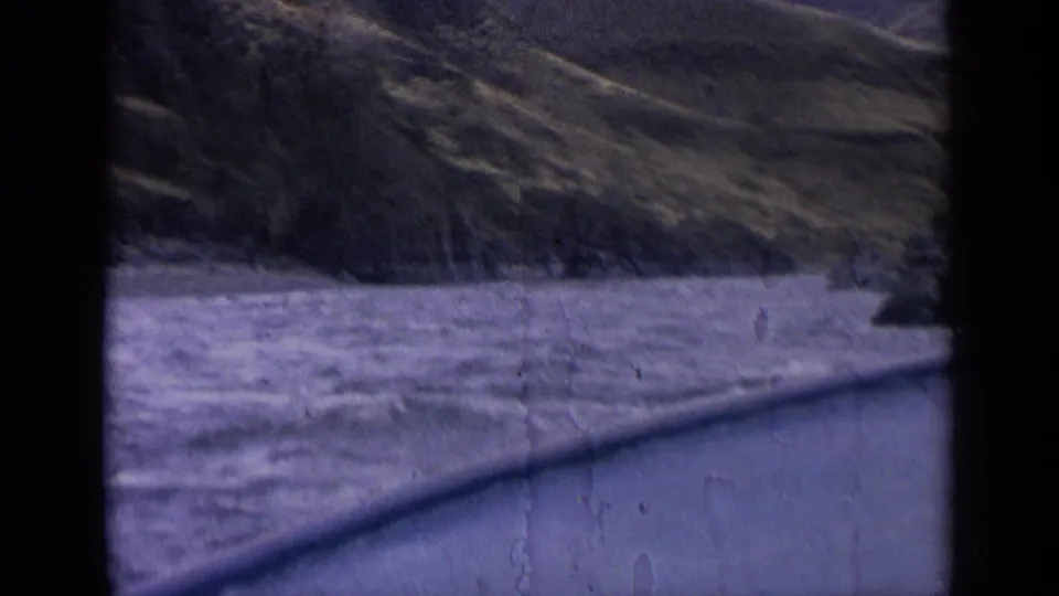 1963 Idaho 8mm frame showing a river below steep rocky hills from a moving roadside perspective.