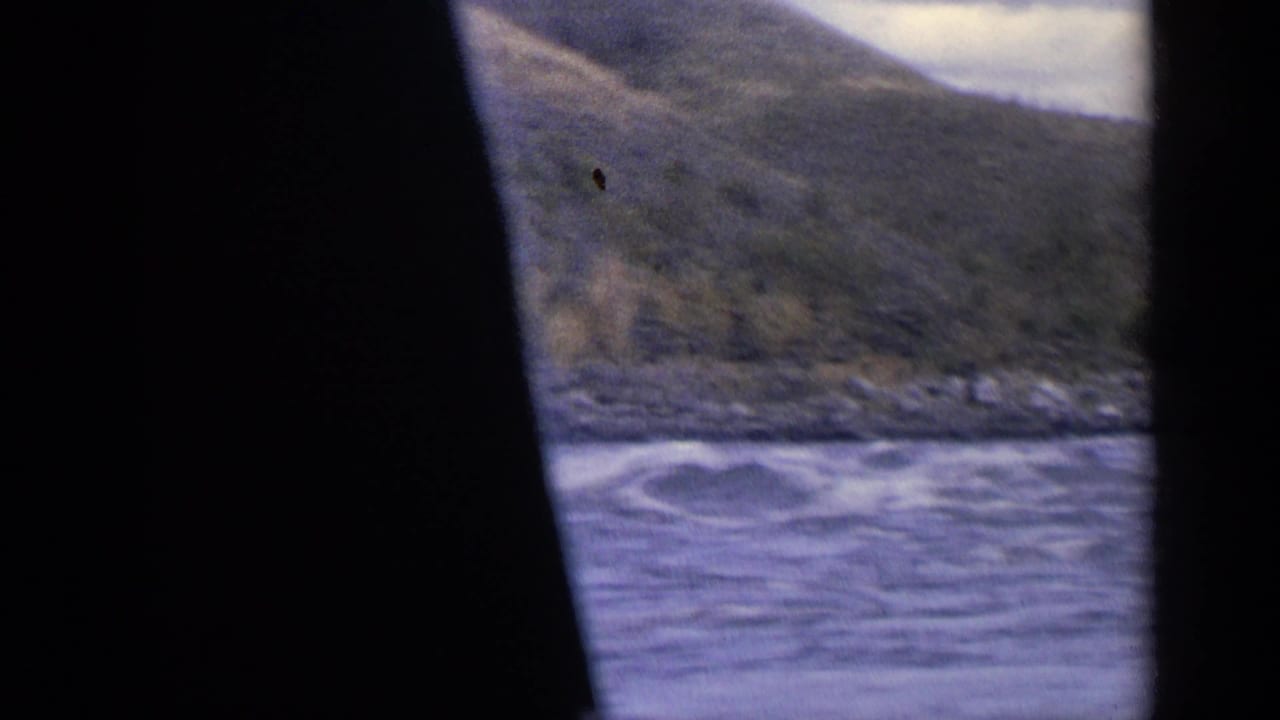 1963 Idaho travel footage shows river water, a rocky shore, and mountain slopes partly obscured by a dark film edge.