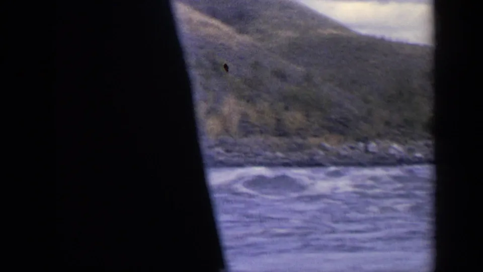1963 Idaho travel footage shows river water, a rocky shore, and mountain slopes partly obscured by a dark film edge.