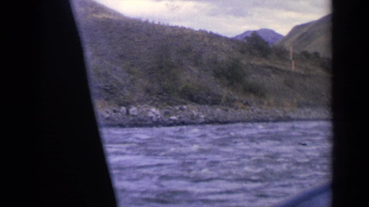 1963 Idaho 8mm frame looking across a river toward rocky bank, wooded hills, and distant mountain shapes.