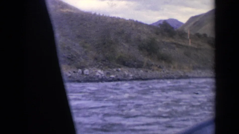 1963 Idaho 8mm frame looking across a river toward rocky bank, wooded hills, and distant mountain shapes.