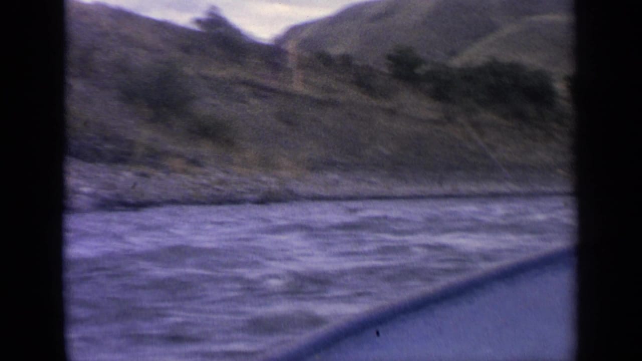 1963 Idaho home-movie frame with choppy river water in the foreground and rugged hills along the bank.
