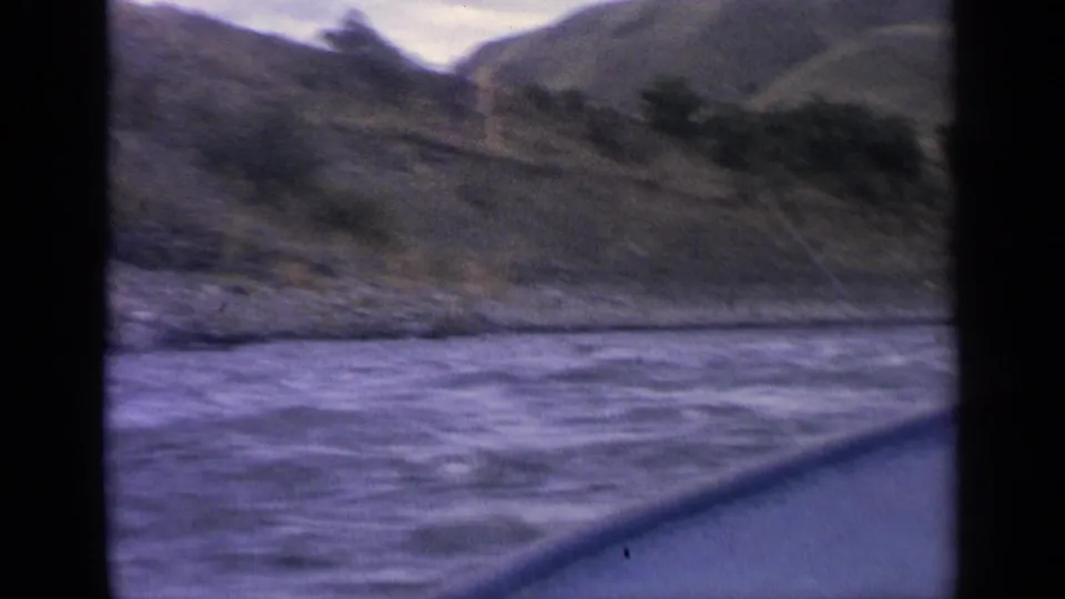 1963 Idaho home-movie frame with choppy river water in the foreground and rugged hills along the bank.