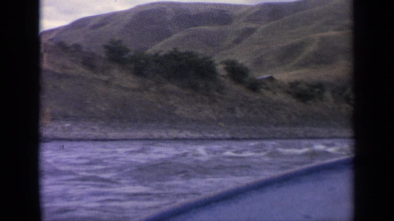Boat passenger view in 1963 Idaho showing rippled river water, brushy shoreline, and rounded hills.