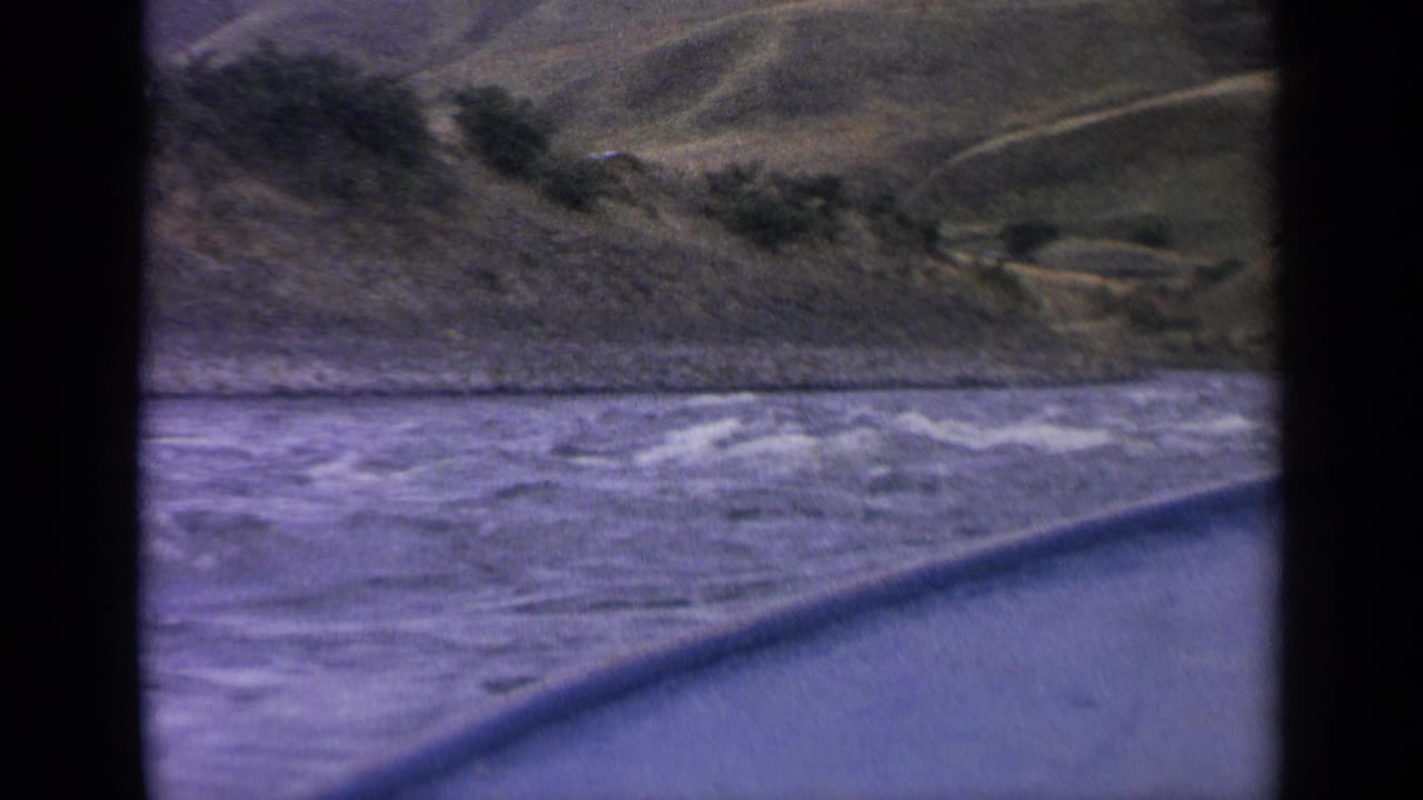 1963 Idaho 8mm frame of a choppy river beside the boat, with sloped hills and scattered shrubs beyond.