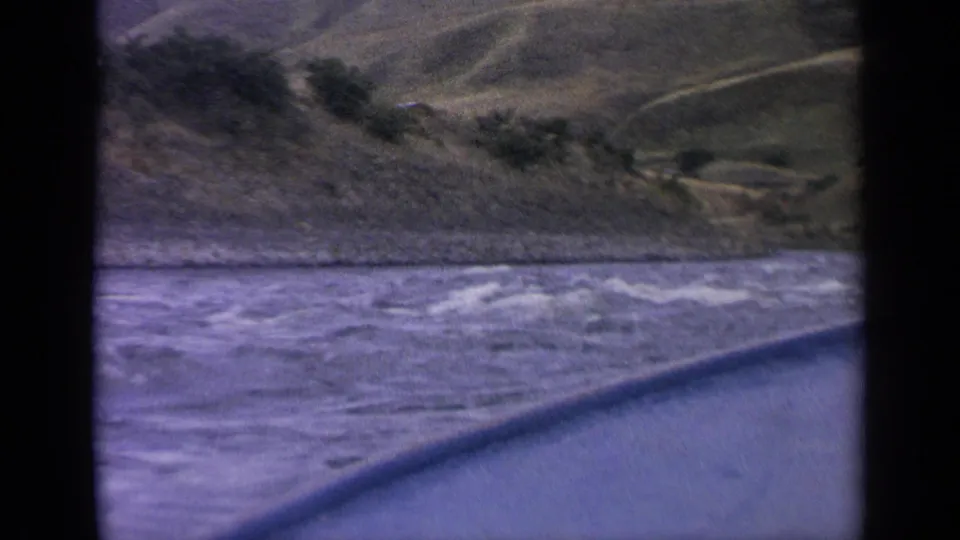 1963 Idaho 8mm frame of a choppy river beside the boat, with sloped hills and scattered shrubs beyond.