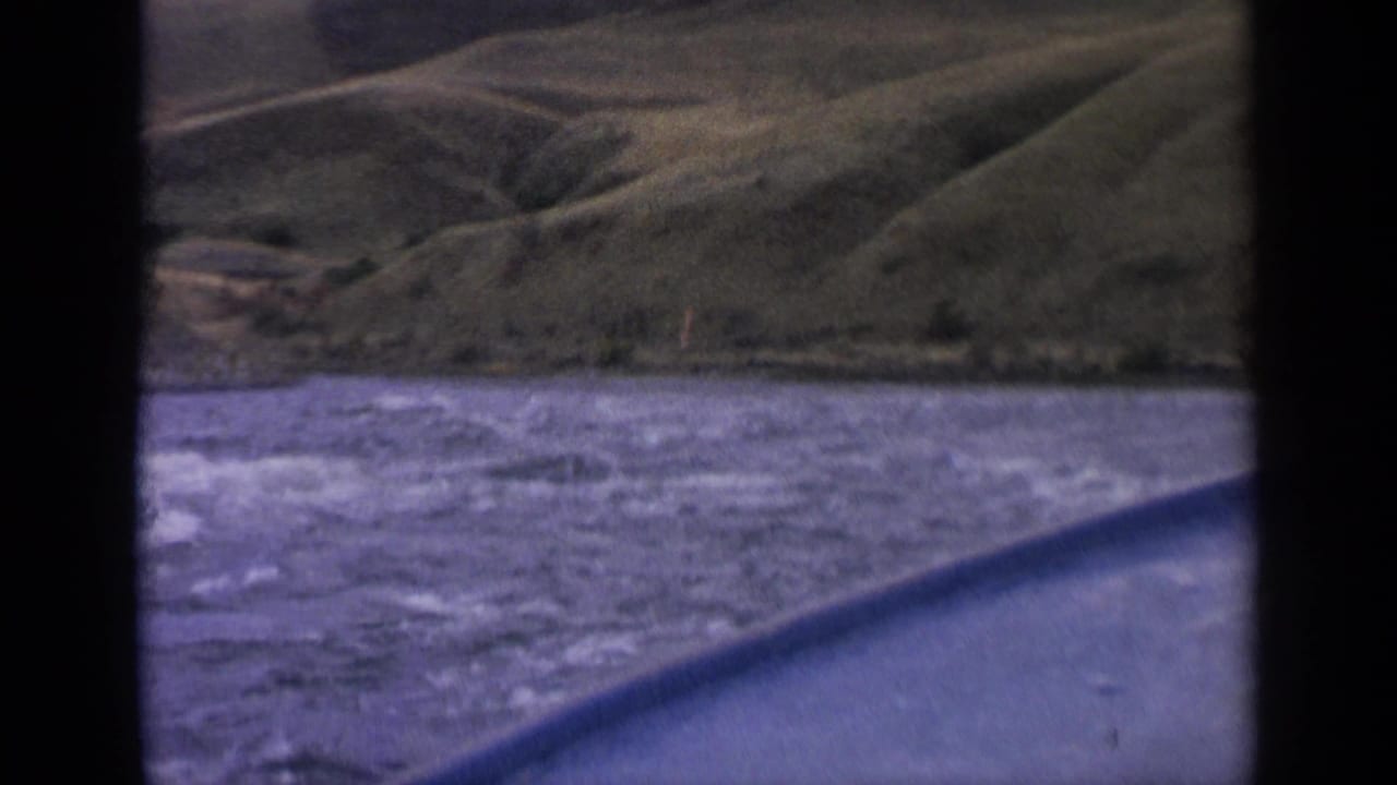 1963 Idaho 8mm frame with rough river water in the foreground and rounded green hills beyond a brush-lined shoreline.