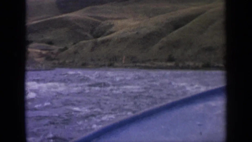 1963 Idaho 8mm frame with rough river water in the foreground and rounded green hills beyond a brush-lined shoreline.