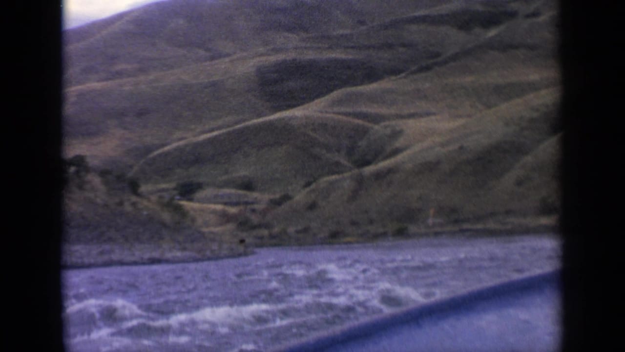 1963 Idaho home-movie frame showing rippled river water from a moving boat, with a green hillside and shallow valley forms beyond the shore.