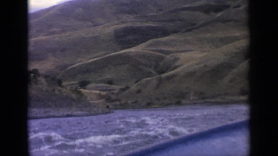 1963 Idaho home-movie frame showing rippled river water from a moving boat, with a green hillside and shallow valley forms beyond the shore.