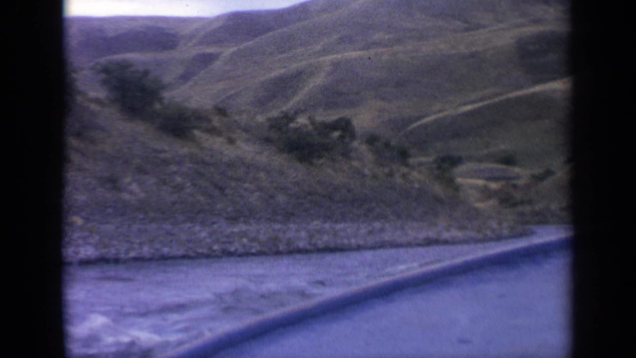 8mm Idaho river-travel frame showing a low boat edge, rippled water, a rocky bank, and green-brown hills in the distance.