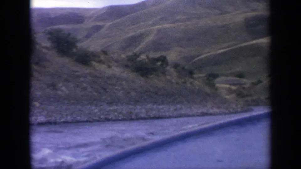 8mm Idaho river-travel frame showing a low boat edge, rippled water, a rocky bank, and green-brown hills in the distance.