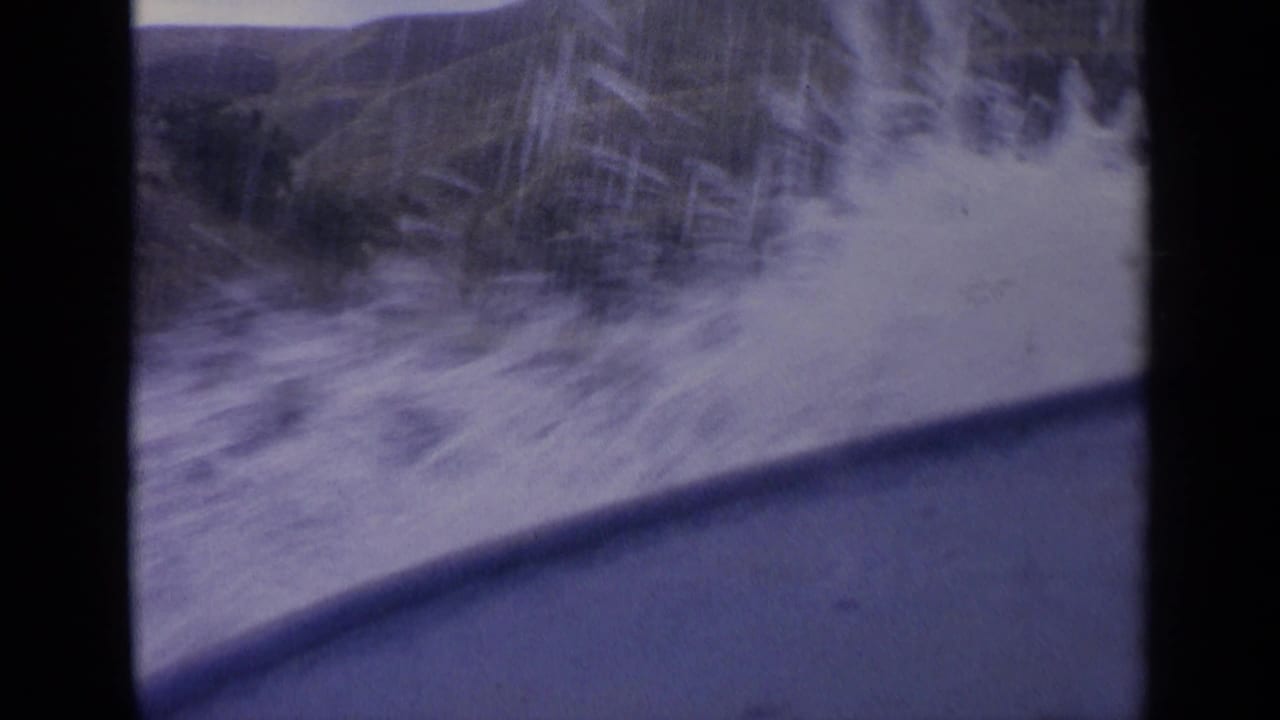 Final 1963 Idaho 8mm frame with heavy white spray beside the boat, soft motion blur, and hillside terrain behind the river.