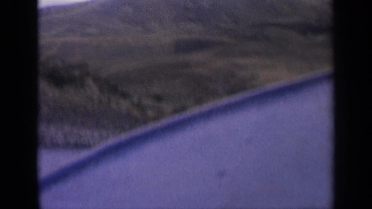 1963 Idaho 8mm road-view frame showing water, dry hillside, and dark vehicle-window borders during travel.