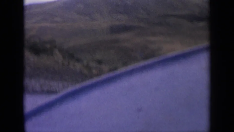 1963 Idaho 8mm road-view frame showing water, dry hillside, and dark vehicle-window borders during travel.