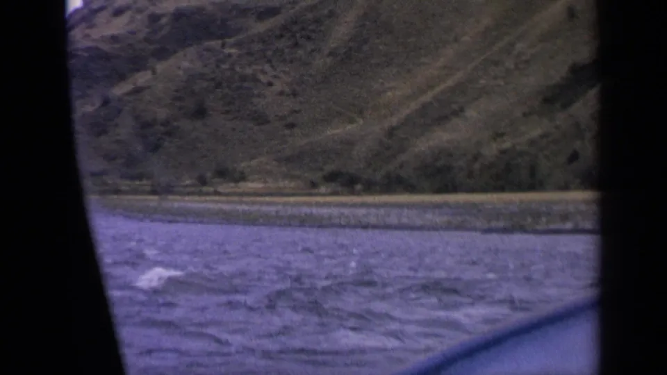1963 Idaho home-movie frame with choppy water, gravel bank, and steep dry hills seen from a moving car.