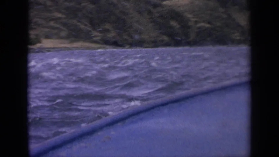 1963 Idaho 8mm still with lake waves in the foreground, a boat edge at bottom, and a green-brown hillside beyond.