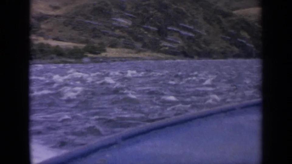 1963 Idaho home-movie frame showing textured hills beyond blue lake water, with the boat edge visible in the foreground.